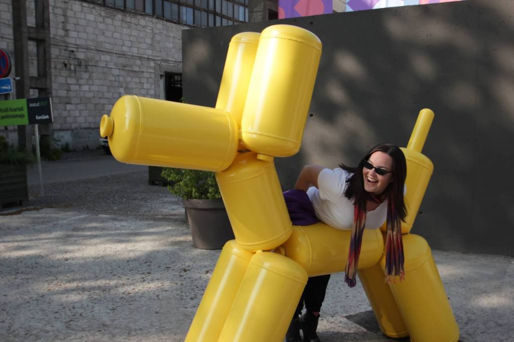 The author Hannah Brown smiling and laughing as she poses with a yellow dog sculpture - representing her embodiment of nonconformity